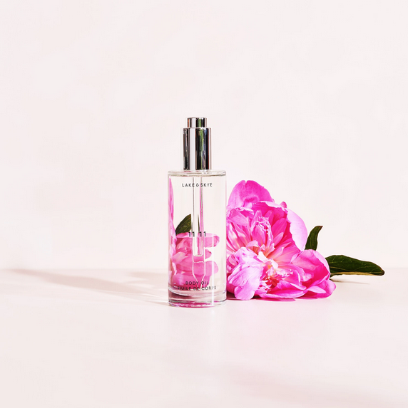 3.4oz bottle of Lake and Skye 11 11 body oil adjacent / infront of pink hibiscus-looking flower